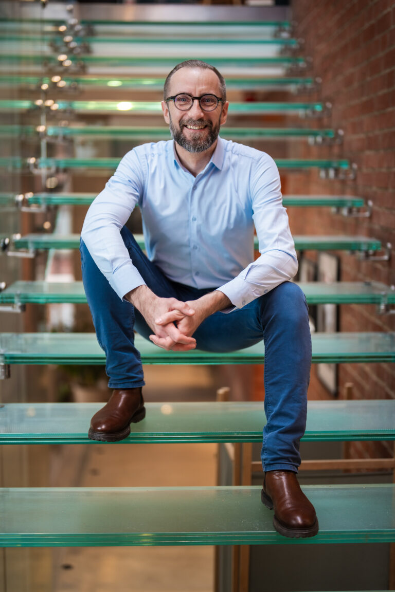 Jakub Dejewski, CEO of IVF Media Ltd., smiling while sitting on glass stairs, wearing a light blue shirt, jeans, and leather boots.