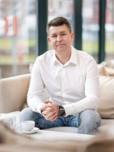 Aleksander Wiecki, CMO of IVFMedia Ltd., smiling in a professional portrait, sitting on a cream sofa wearing a white shirt and jeans